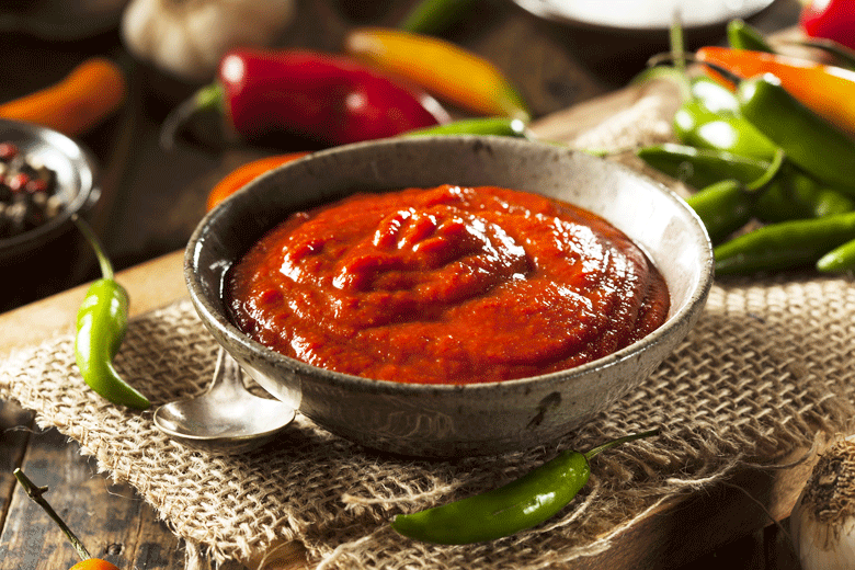 A bowl of homemade Sriracha sauce on a wooden table surrounded by green and red Chillies 